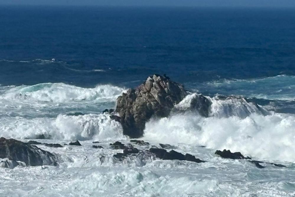 Powerful ocean waves crash against jagged offshore rocks, sending white spray and churning foam across the dark blue water under an open horizon.